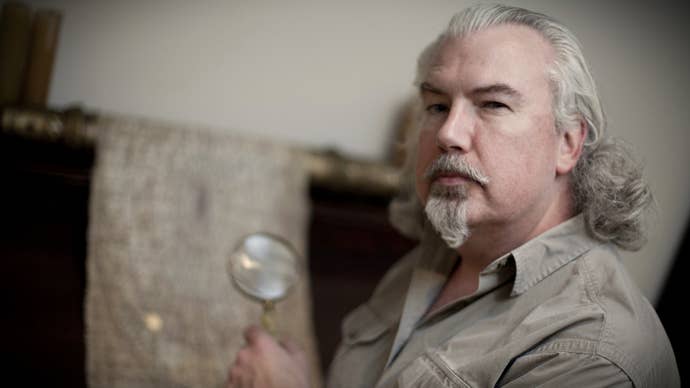 A photograph of a white man with a pointed, Van Dyke white beard, and long white hair neatly swept back over his head, holding a magnifying glass up to some ancient document or other in the background.