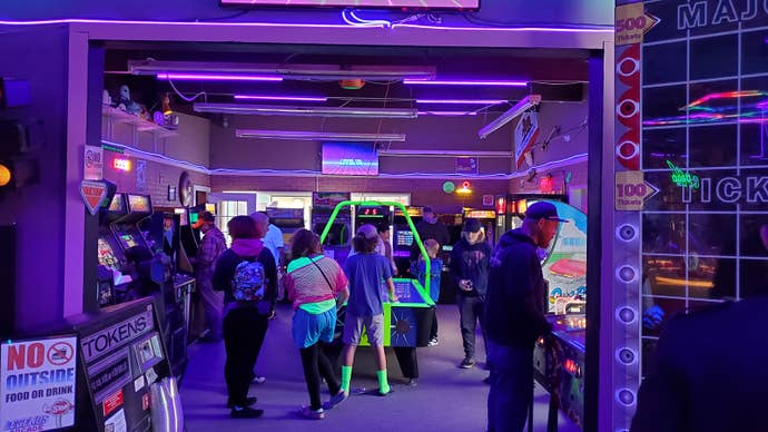The inside of Legends Arcade. People crowd around arcade cabinets and an air hockey table in an ultra violet, purple and neon environment.