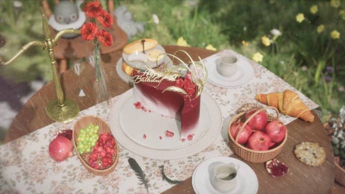 A close-up of the surface of a round wooden table placed in a field. On the table is a floral runner and baskets of grapes and other fruits, teacups, a croissant, and a couple of black feathers. The centrepiece, however, is a striking crescent-shaped red cake with elabourate gold feather and red gem toppers and Happy Birthday written in gold.