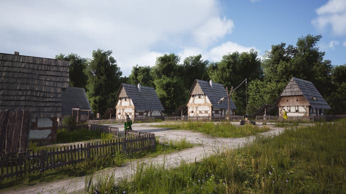 A cluster of thatched and daubed medieval houses stand in a countryside of deep green grass and blue but cloudy skies.