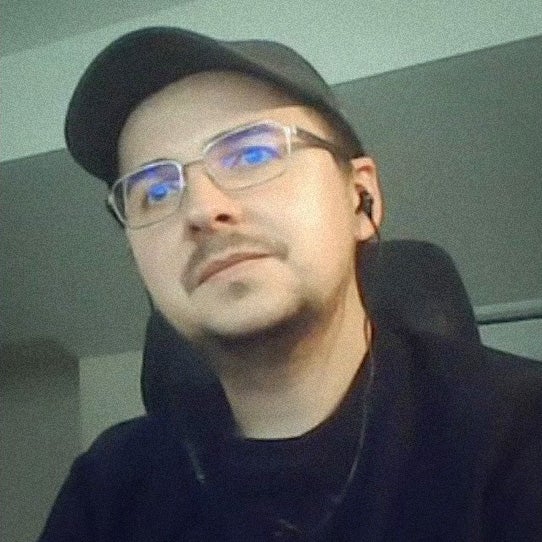 A photo of a white man with rectangular glasses, baseball cap, and short goatee. They sit in an office, looking slightly off to the side from the camera. It's Maxim Verehin.