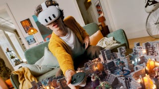 A promotional image for Meta Quest 3 showing a man wearing a headset while sat on the sofa in his living room. He leans forward holding a controller, his hand hovering above a virtual fantasy dungeon superimposed on the table.