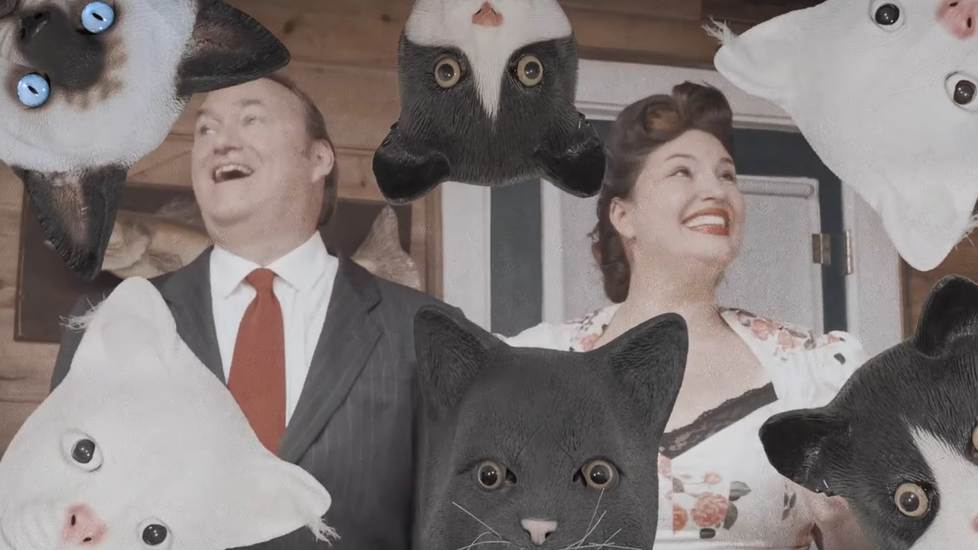 A smiling, happy, '60s-esque couple are smiling surrounded by people wearing slightly realistic cat masks in a trailer for Mewgenics. - 1