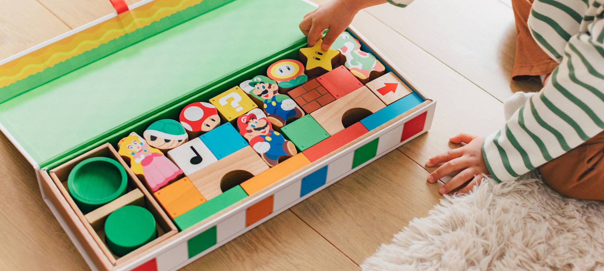 Promotional photo showing a young child reaching into a large box containing Mario-themed wooden blocks.
