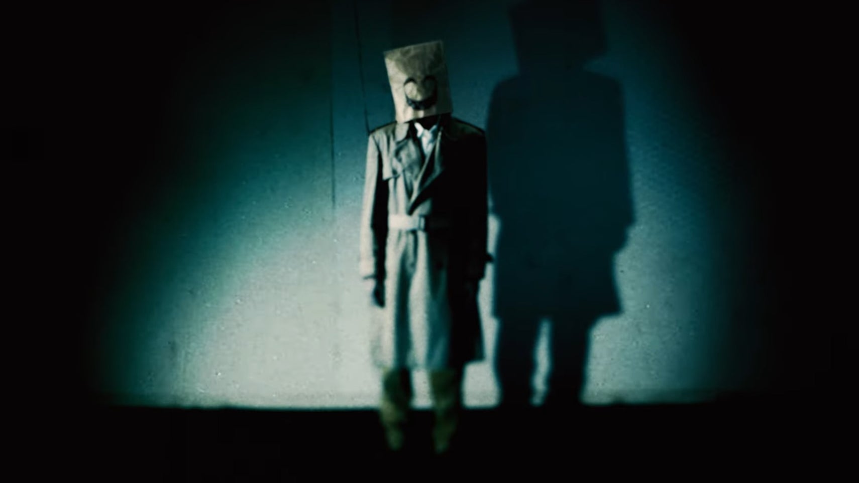 A man in a trench coat and paper bag with a creepy smiling face on it is stood ina dimly lit room, the image is covered in a film grain effect. - 1