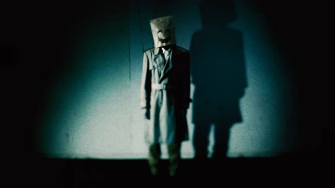 A man in a trench coat and paper bag with a creepy smiling face on it is stood ina dimly lit room, the image is covered in a film grain effect.