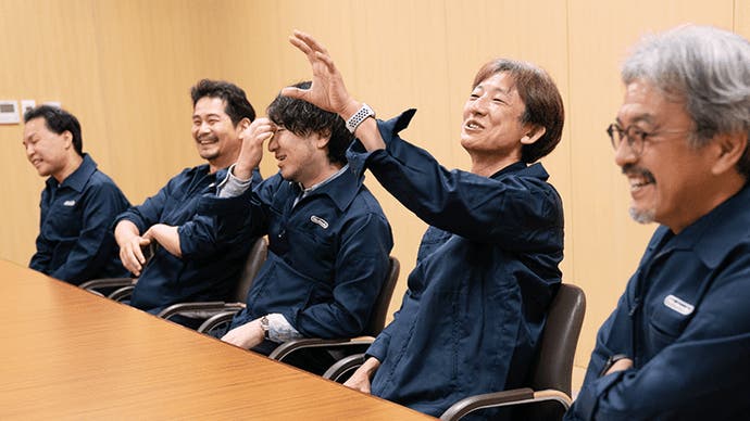 Zelda developers in blue jumpsuits at a table laugh during one's explanation of something. From left to right: Hajime Wakai, Satoru Takizawa, Takuhiro Dohta, Hidemaro Fijubayashi, Eiji Aonuma