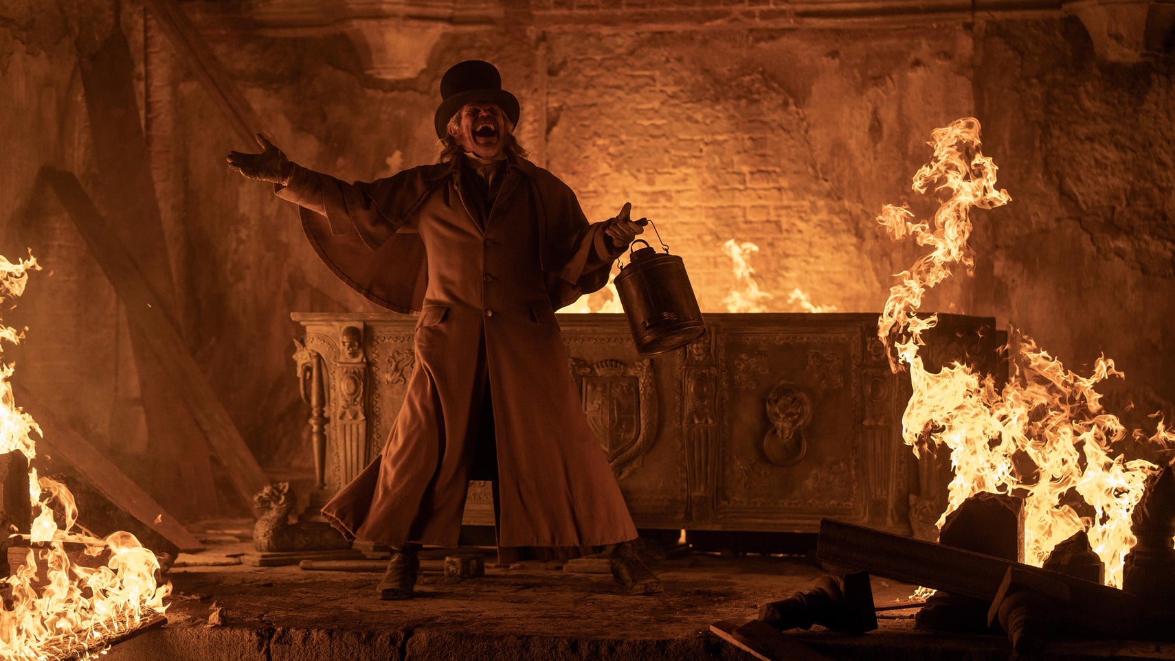 Willem Dafeo in 2024's Nosferatu, he's dressed in old attire, holding some kind of metal container stood in what looks like a mausoleum, which is partially on fire. - 1