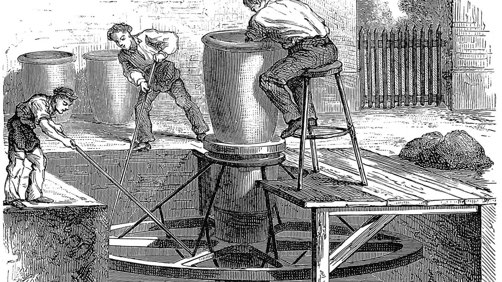 An old black and white vintage illustration of a potter sitting on a three-legged stool shaping a tall pot, using a large spoked wooden wheel whose lower part is lodged below the floor level of the workshop and which is operated by two boys equipped with long sticks. - 1