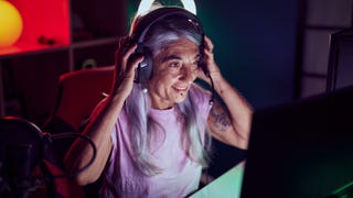 A woman with long grey hair pulls on a gaming headset in front of a glowing screen.