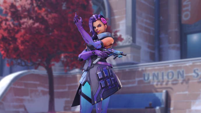 Sombra, a hero in Overwatch 2, poses outside a building in front of the camera, with a red-leaved tree behind her on the left.