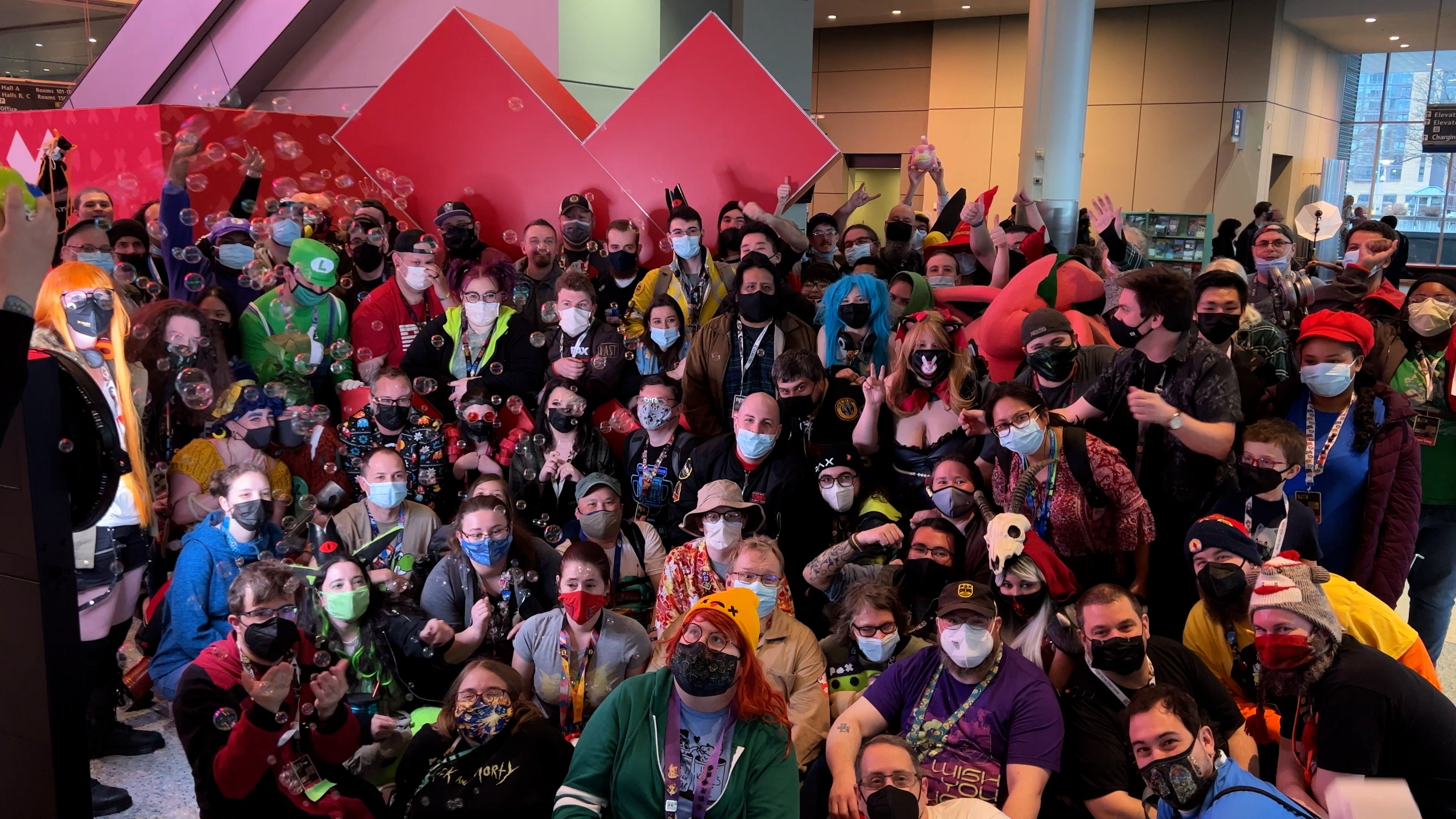 A large group of people, some wearing costumes of their favourite video game characters, gather around a large PAX logo to take a photograph together.