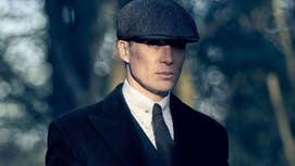 Cillian Murphy as Tommy Shelby in Peaky Blinders, he's stood in a plain suit and a flat cap with a stern expression on his face in the woods.
