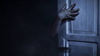 A hand in the dark reaches around an open door, and there's something not quite right about it. A scary image.