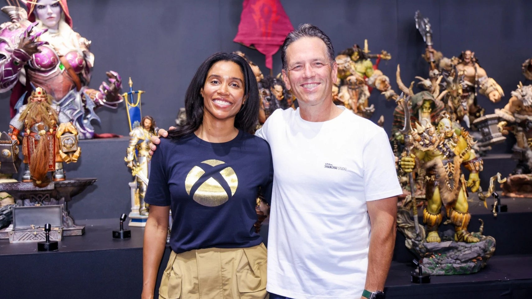 Former Xbox and Microsoft gaming execs Sarah Bond and Phil Spencer stood army in arm at a convention in China. Many Warcraft-related figures are behind them.