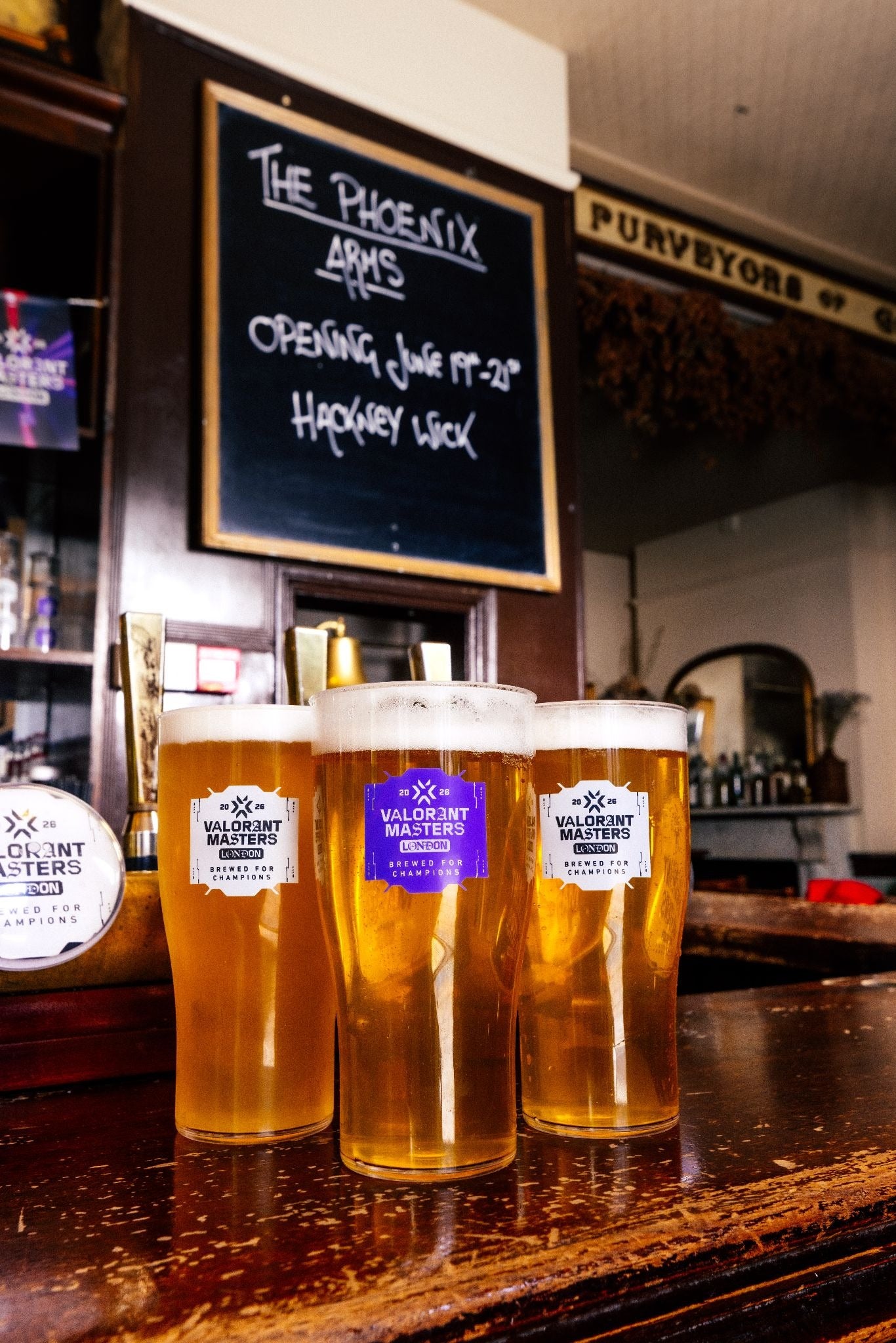 Three full pint glasses stand on a bar, waiting to be demolished. They have Valorant logos on the glasses. There are no hands in sight.