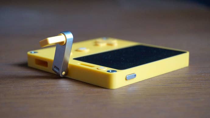 The Playdate lies on a wooden table, showing the crank in an upright position and the top lock button