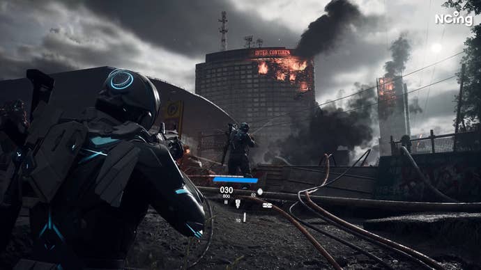 A sci-fi man points his gun at another sci-fi man as buildings burn in a grey modern world.