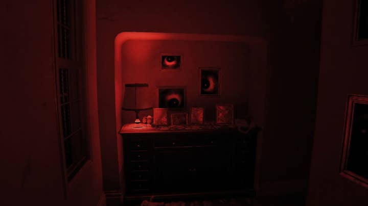 P.T. screen of a desk in a hallway lit only by a red light coming from an unclear source. There are three framed pictures on the wall, but each one is an extreme close-up of an eye looking in different directions