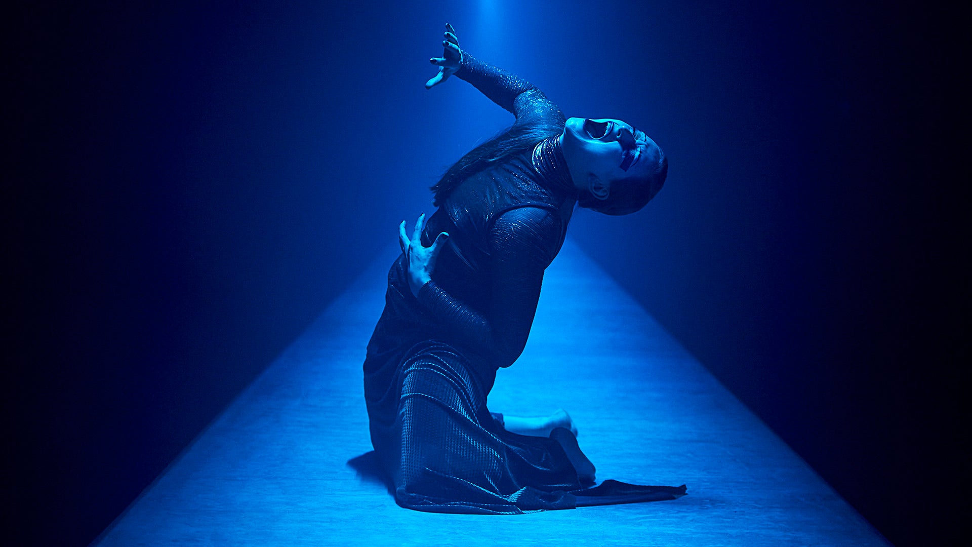 A feminine person doubled back in agony on a seemingly endless table, bathed in blue light.