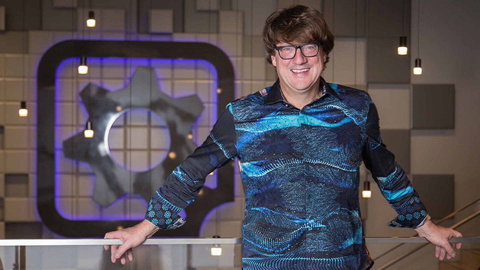 Randy Pitchford, a middle-aged man with glasses and a long fringe haircut, is stood in front of a railing and the Gearbox logo. - 1