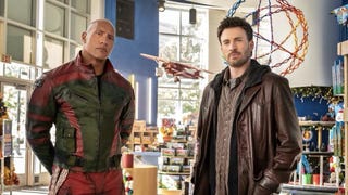 Dwayne Johnson, wearing black and red leather, is stood with Chris Evans, in a brown outfit and leather jacket, in a toy shop in a promotional image for Red One.
