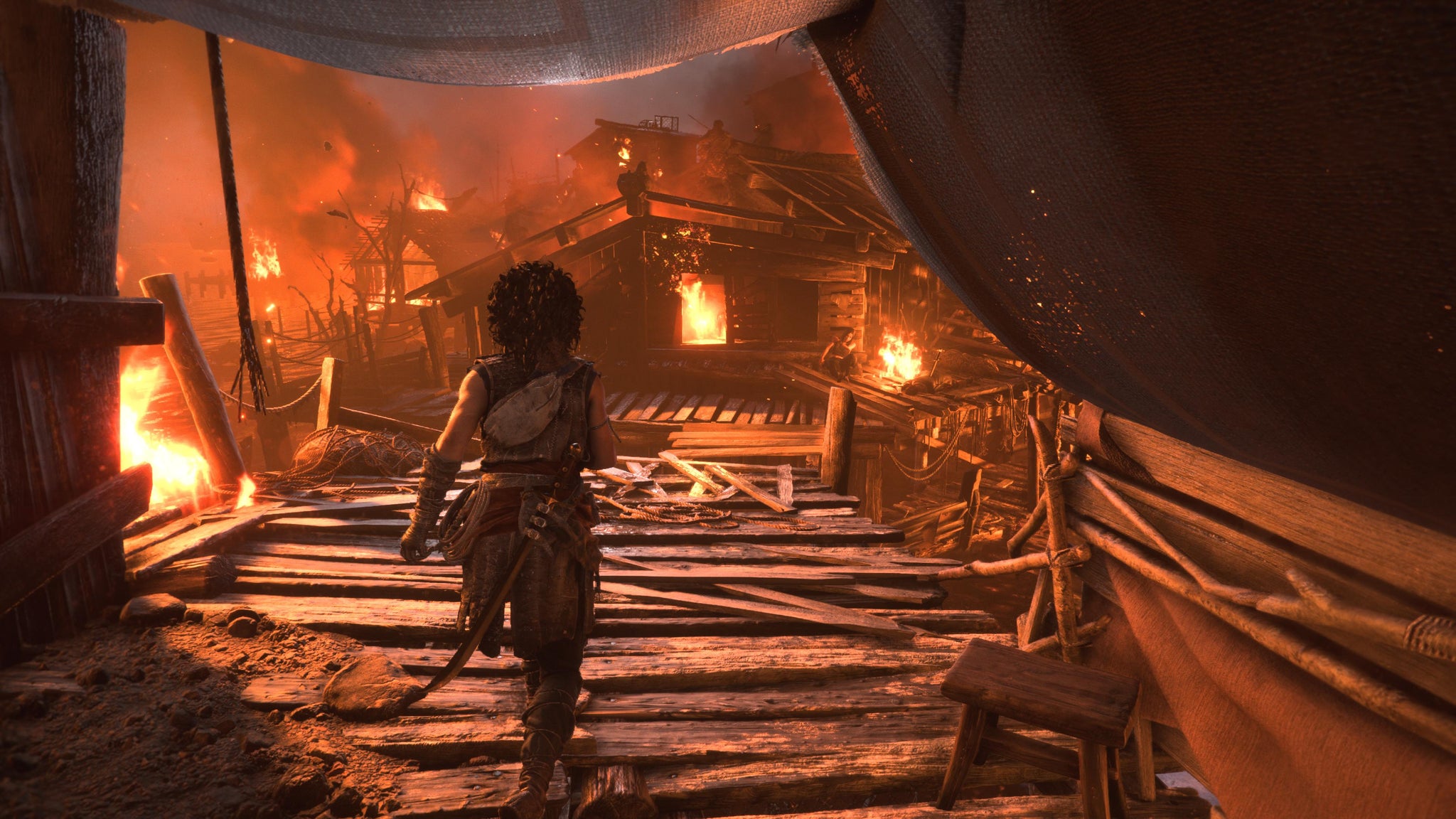 Resonance: A Plague Tale Legacy screenshot showing a traditional video game over-the-shoulder shot of player character Sophia advancing towards a series of wooden cabins that are aflame