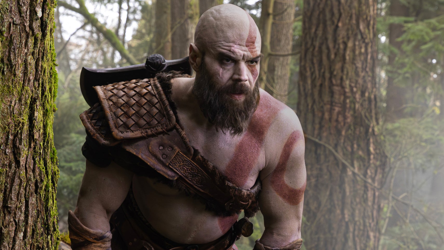 A photograph of actor Ryan Hurst as Kratos in God of War. He is topless, muscled, and wears a leather harness across his torso. He has red stripes on him.