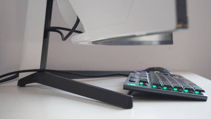 A photo of the Samsung Odyssey G9 gaming monitor's stand from a side-on view, with a keyboard and mouse