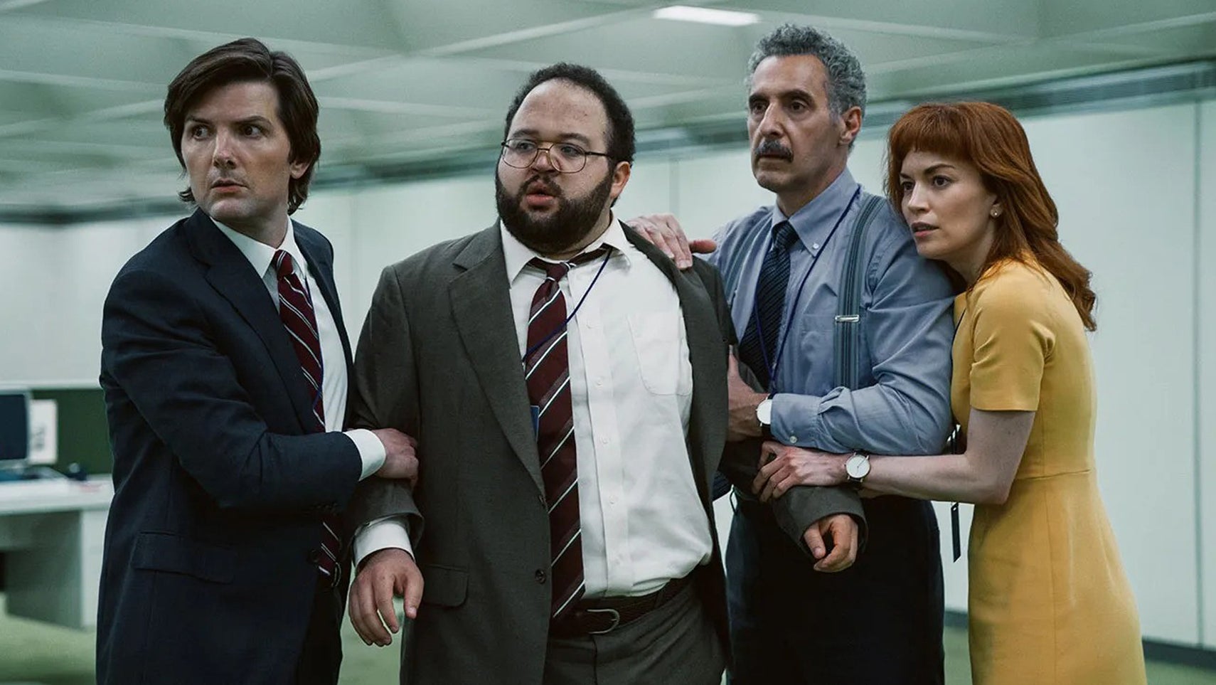 The main cast of Severance, including Adam Scott, Zack Cherry, John Turturro, and Britt Lower, hudled together in an office space looking at something offscreen. - 1