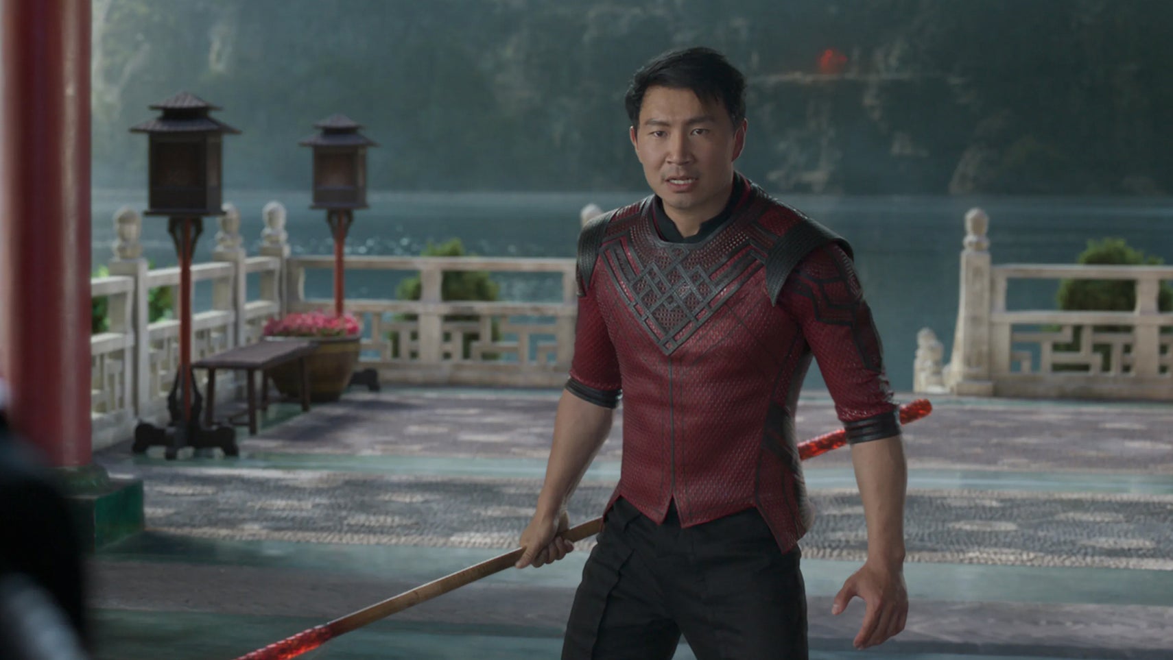 Shang-Chi is stood on some decking overlooking a lake, a staff in his hands. - 1