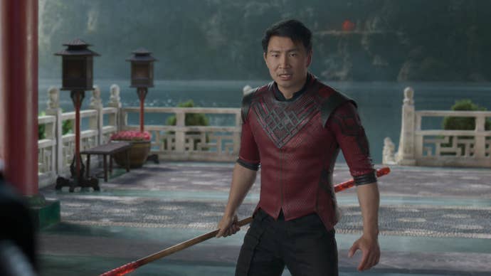 Shang-Chi is stood on some decking overlooking a lake, a staff in his hands.