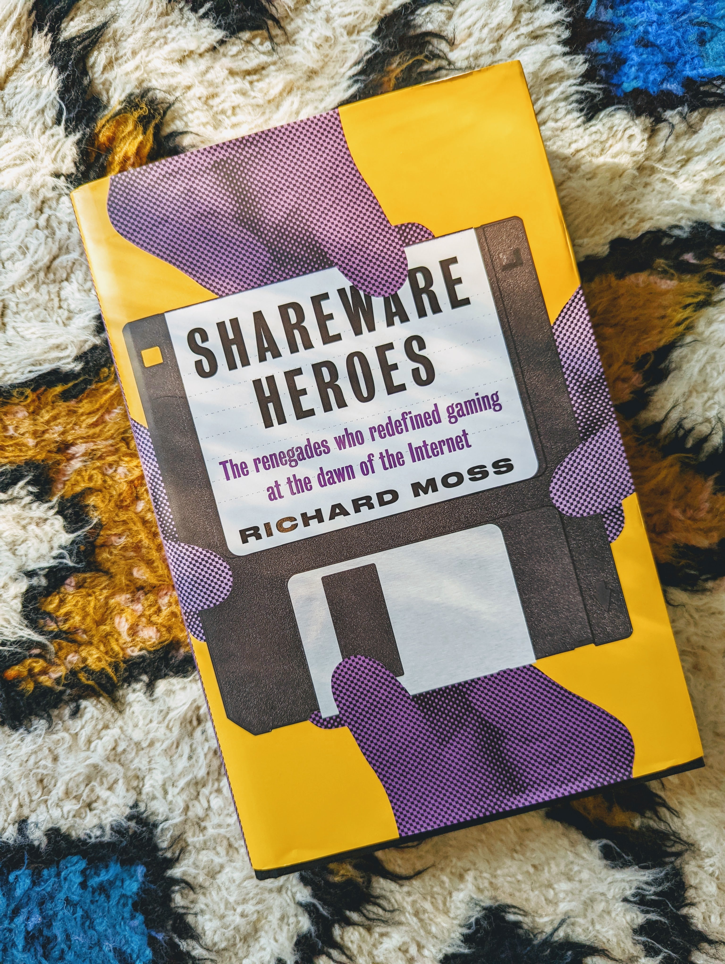 The Shareware Heroes book, pictured on Bertie&rsquo;s wonderful multi-coloured rug. The book cover has four purple hands pulling at a floppy disk bearing the name of the book and the author Richard Ross. The rest of the book is coloured yellow.