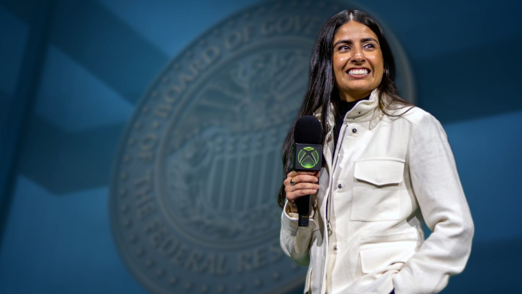 Asha Sharma, holding an Xbox microphone, in front of an emblem of the US Federal Reserve