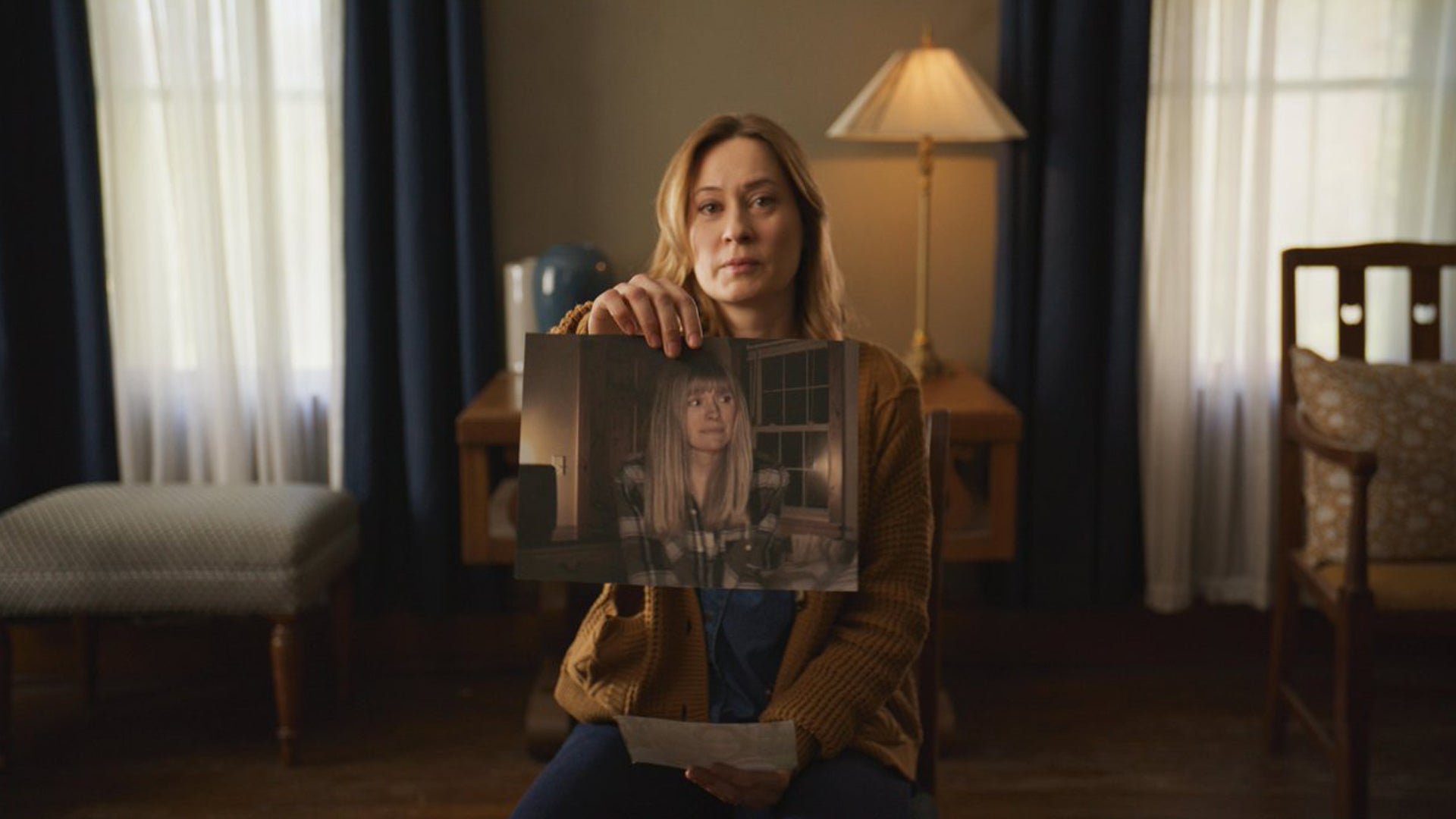 A woman is sat on a chair in a bedroom holding up a photograph of another woman.