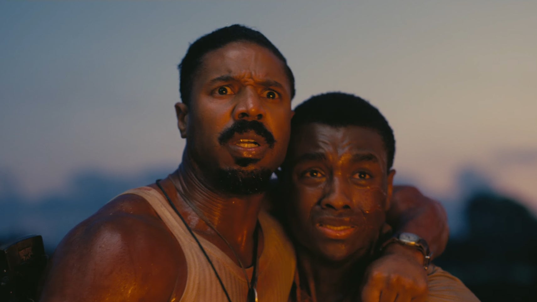 Two men, one holding the other close to him, look terrified of something offscreen, a warm firey glow hitting their faces in Sinners. - 1