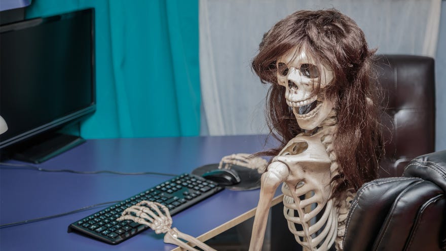 A skeleton with long hair turns to the camera while typing on a keyboard and using a mouse in front of a computer monitor.
