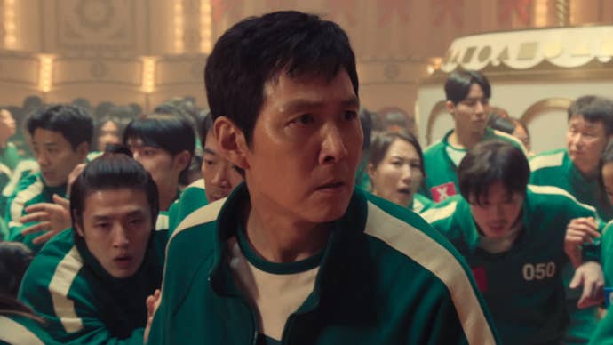 Gi-hun looking concerned as various other Squid Game participants are huddled behind him, also looking worried.