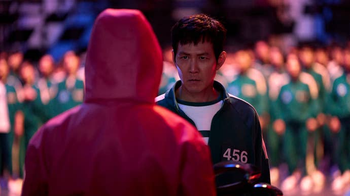 Gi-hun, a middle-aged man, is wearing a green tracksuit, a large number of people in the same outfit out of focus behind him in Squid Game. He is looking at someone in a pinkish jumpsuit.