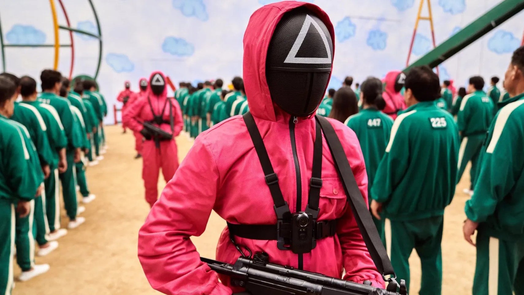 A person in a light red jumpsuit wearing a fencing mask with a white triangle on it in Squid Game. They are holding a gun, others like them lined up next to people in green tracksuits. - 1
