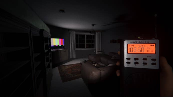 A screenshot of Phasmophobia showing a dimly lit living room viewed in first-person at night. Its furnishing are cast in shadow and the only source of light is a TV screen showing a test signal near a window to the rear of the room. The unseen investigator surveying the scene holds up a small handheld electronic device labelled ‘spirit box’ with two rows of buttons beneath an orange digital display.