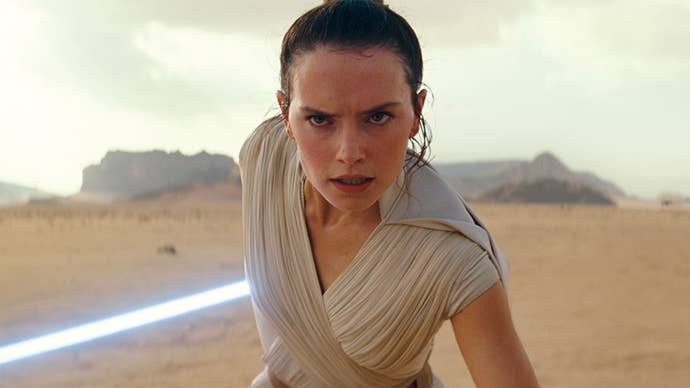 Rey from Star Wars stood in a desert area, a serious expression on her face, lightsabre active in one hand.