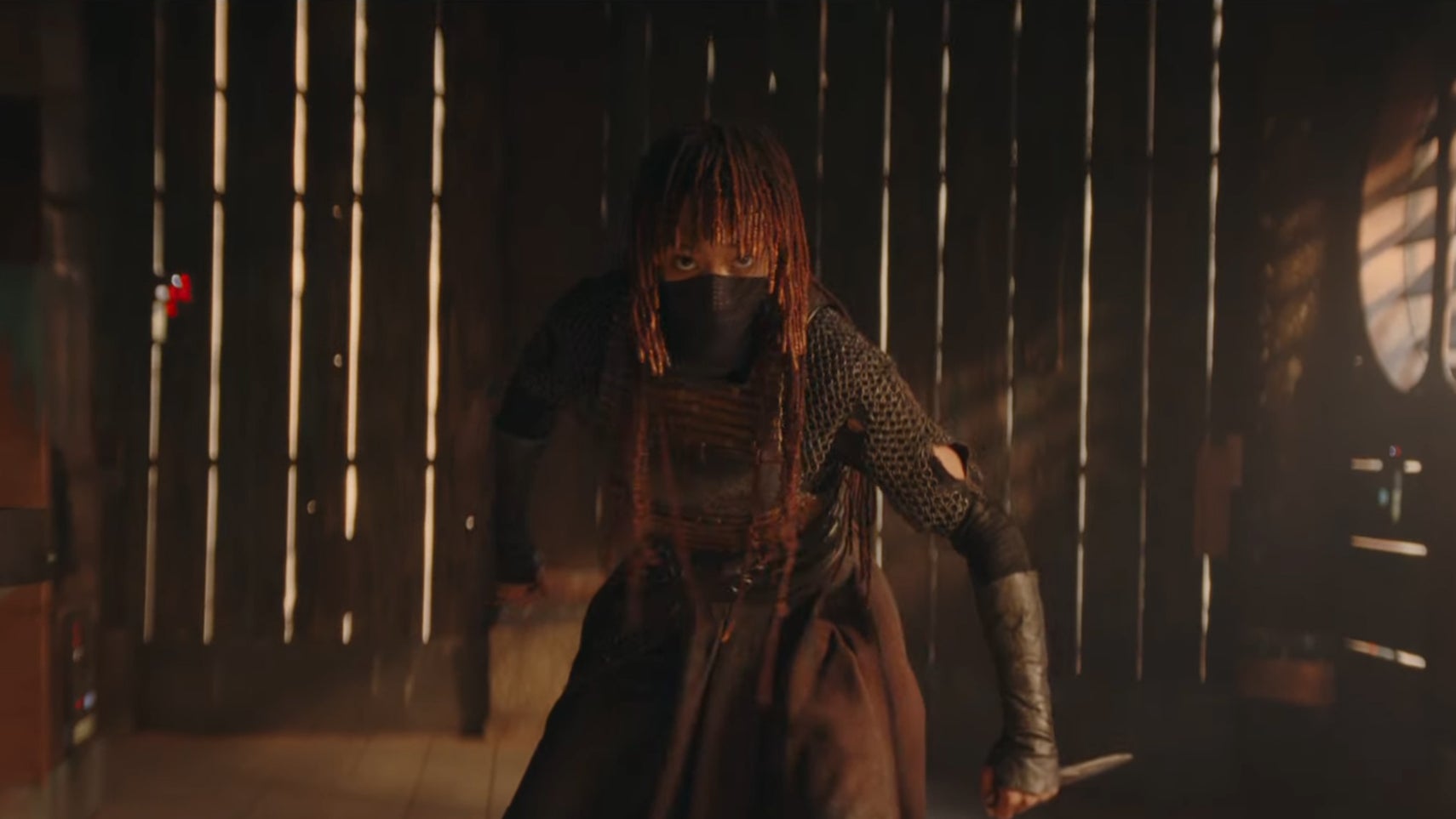 A woman is in a dimly lit bar, she has a mask covering her lower face, holding a knife in one hand in The Acolyte. - 1