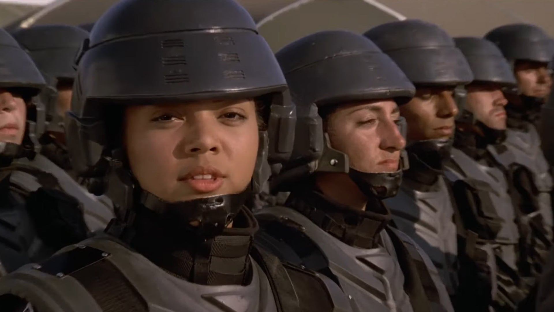 A close-up of a woman soldier, other soldiers lined-up next to her, she&rsquo;s looking into the camera.