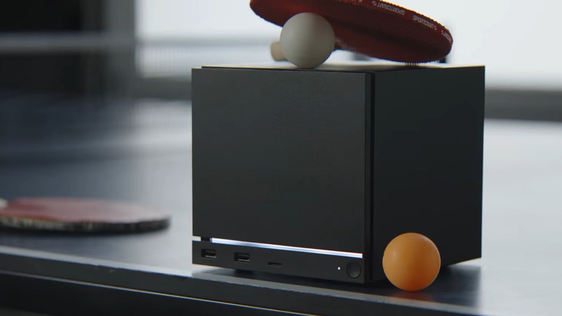 A Steam Machine, which is a cube-shaped, black video game console, is sat on a ping-pong table, a paddle and ball sat on it and next to it. - 1
