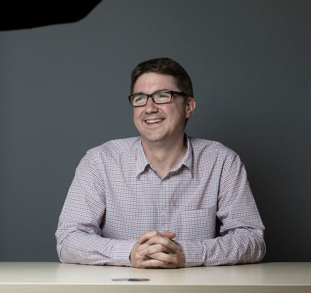 A professional shot of Steve Collins, a man in a checked shirt and no tie, smiling at a desk with hands clasped