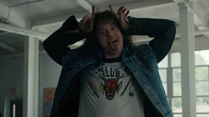 Eddie Munson (Joseph Quinn) mockingly holds his fingers in a “devil horn” shape while sticking his tongue out, making fun of his own satanic reputation. His t-shirt reads “Hellfire Club” and has a picture of a demon’s face on it.