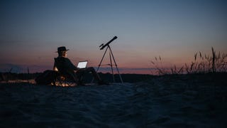 Photo by Pavel Danilyuk of a man sitting in a sandy flat landscape at dusk. He wears a hat and looks at a laptop. A silhouette of a telescope stands next to him