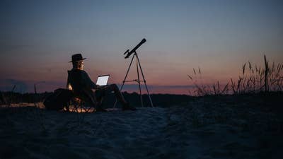Photo by Pavel Danilyuk of a man sitting in a sandy flat landscape at dusk. He wears a hat and looks at a laptop. A silhouette of a telescope stands next to him