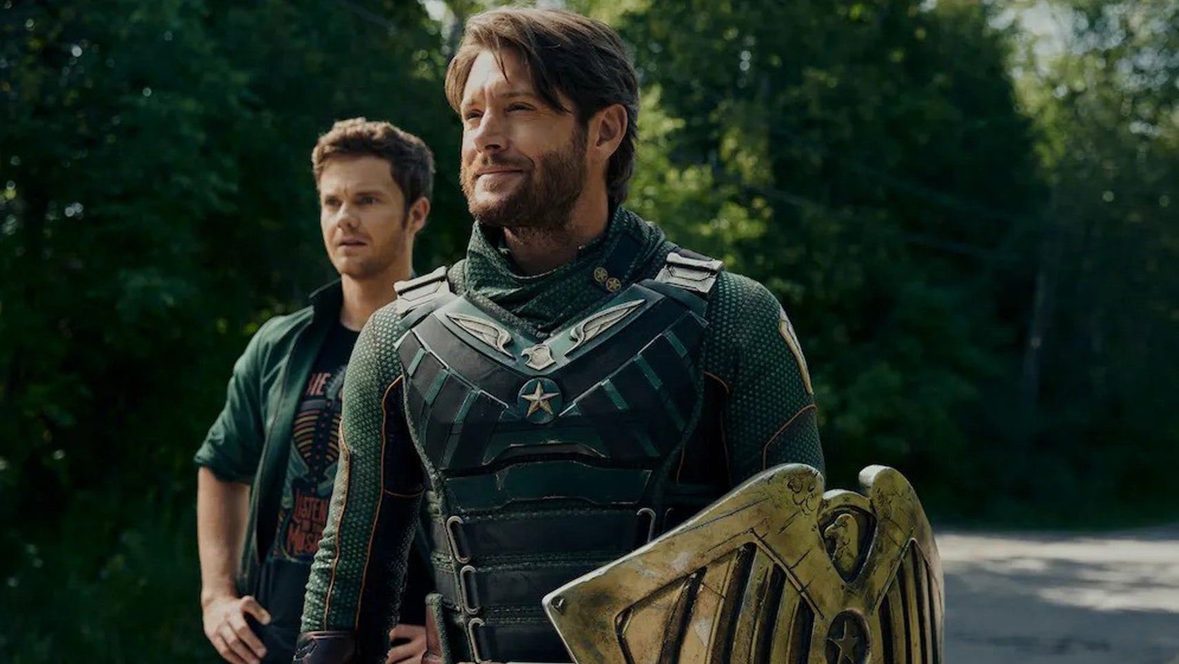 Soldier Boy and Hughie in The Boys, they're both stood on a road in some woods, Soldier Boy stood in his superhero uniform smiling, Hughie looking slightly surprised at something, hands on his hips. - 1